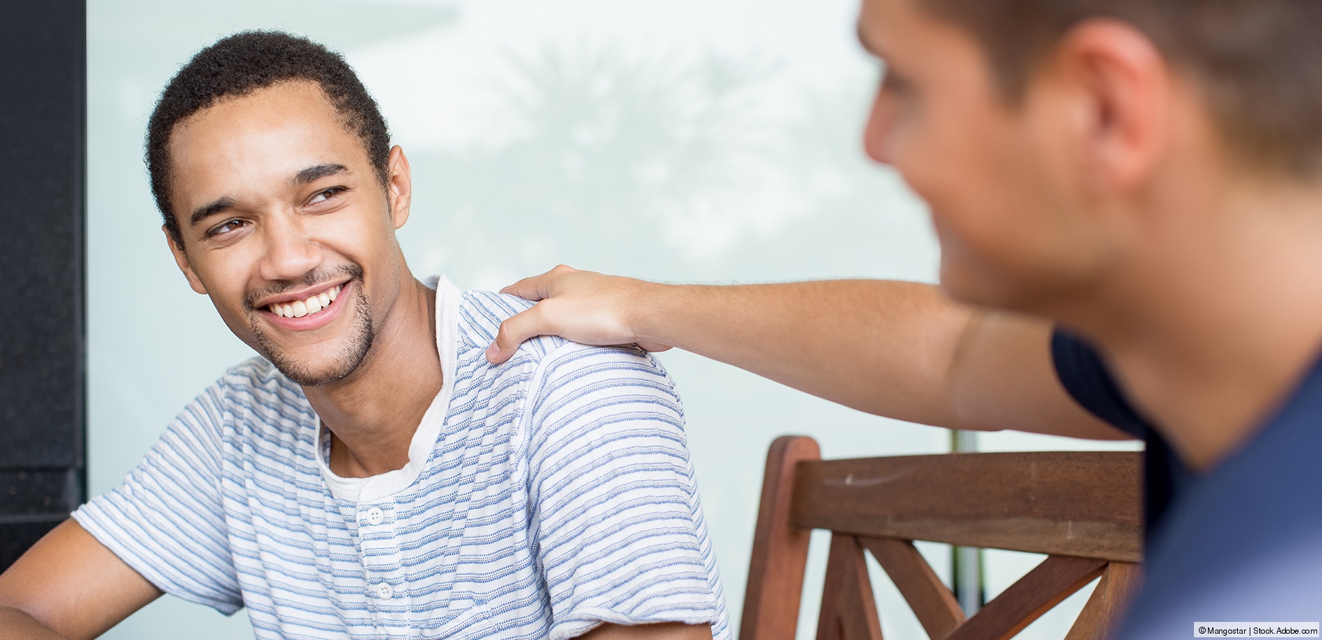 A young, smiling adult has his hand on the shoulder of another smiling young sitting next to him.