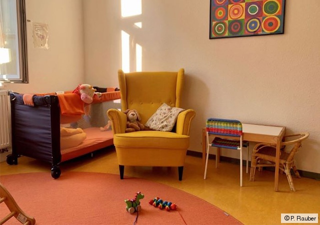 Eltern-Kind-Zimmer, eingerichtet mit Kinderbett, Ohrensessel, Mal-/Spieltisch und Spielzeug