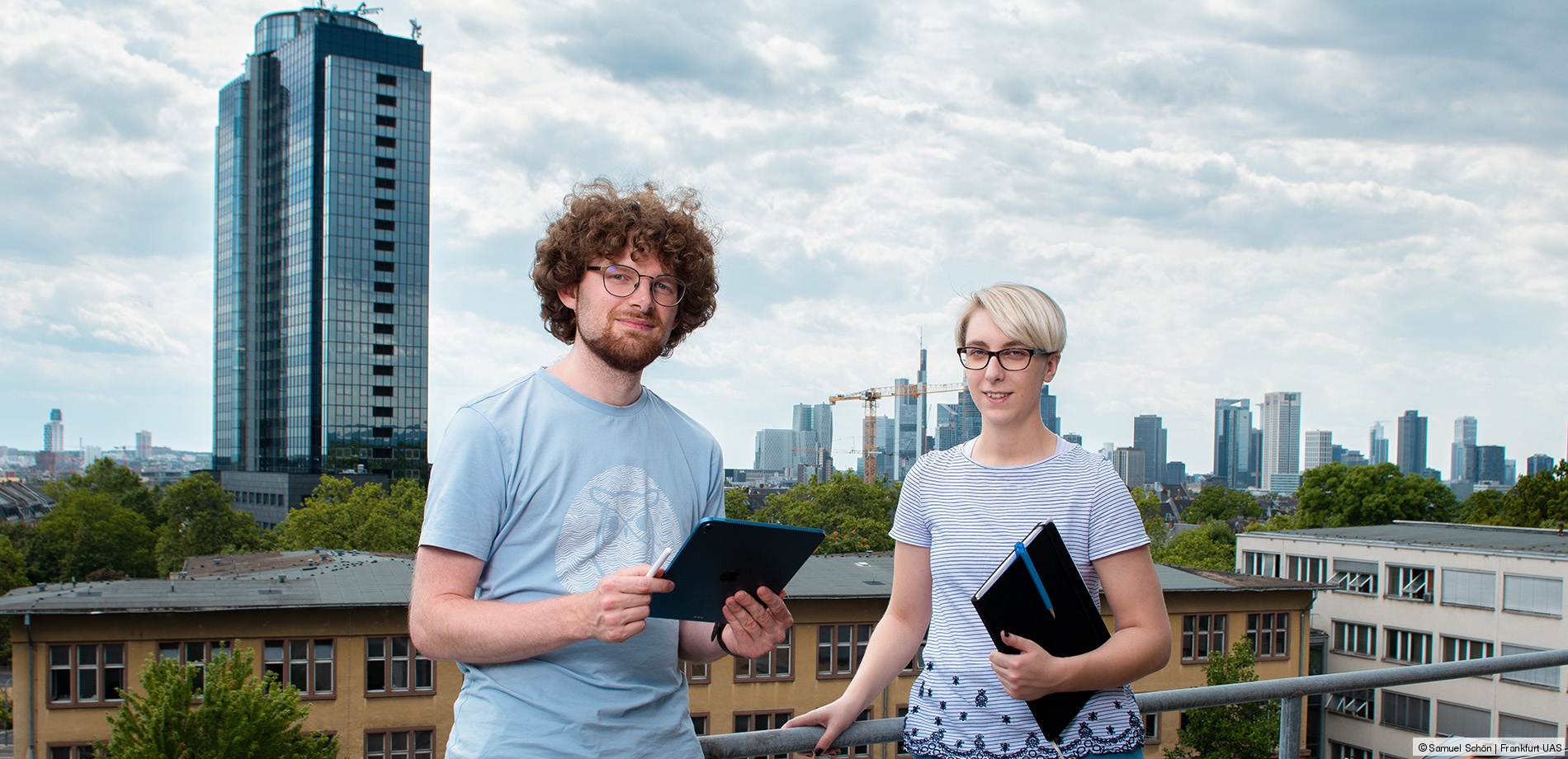 Zwei Personen stehen vor einer Dachbegrenzung, im Hintergrund ist die Frankfurter Skyline zu sehen.