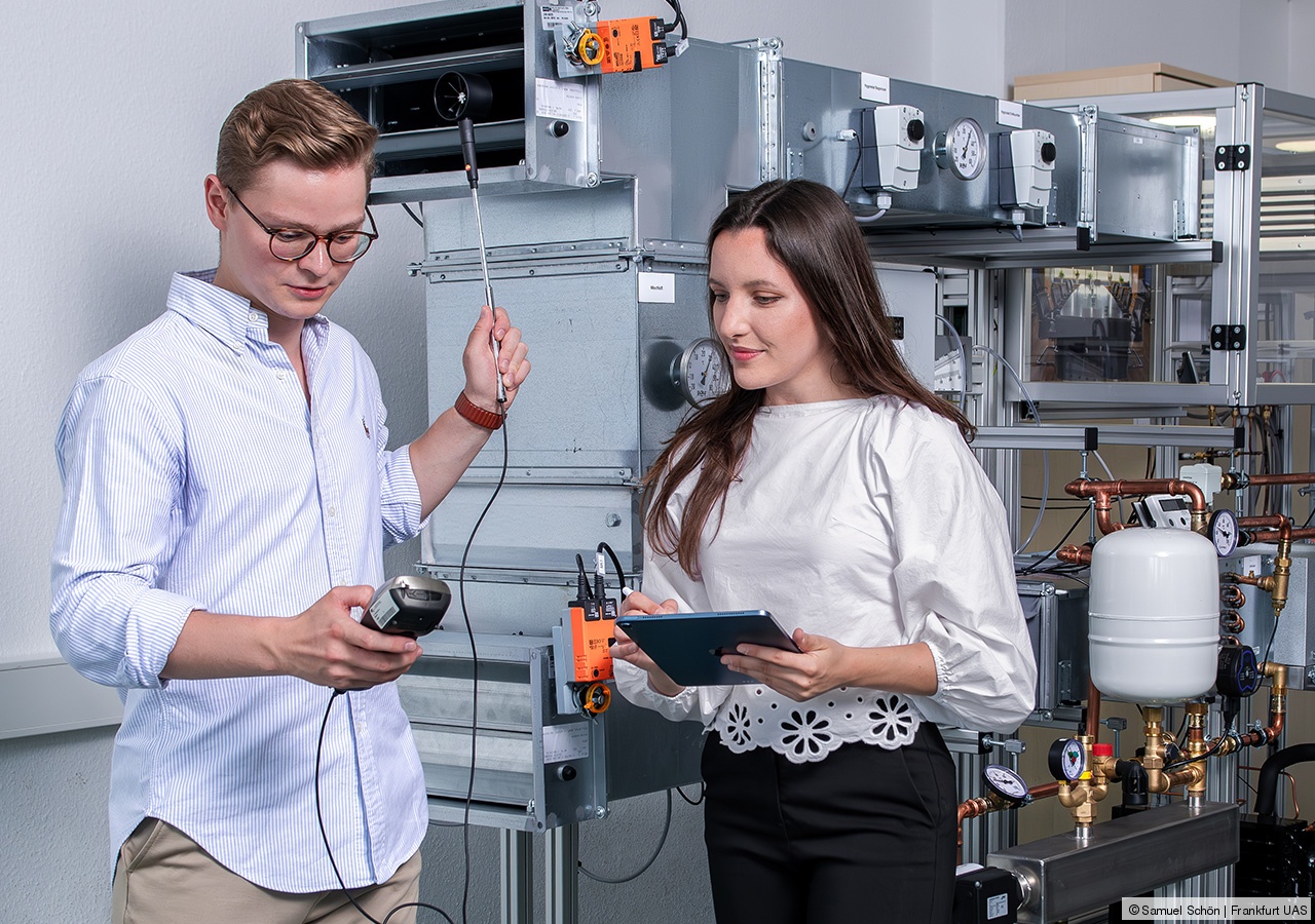 Eine Person hält ein Messgerät und eine kleinen Ventilator in den Händen, eine zweite Person steht daneben, im Hintergrund ist eine Klimatechnikanlage zu sehen.