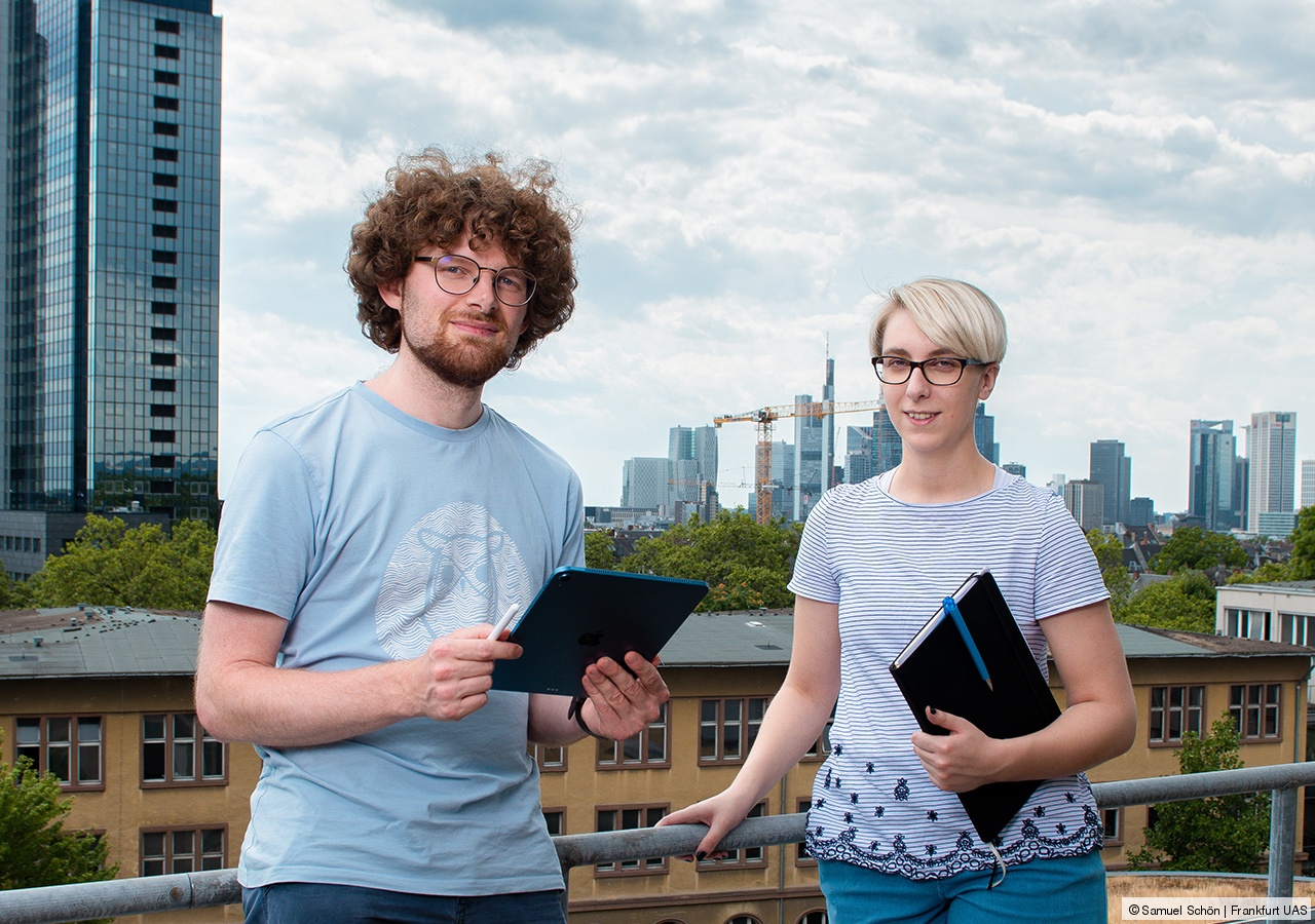 Zwei Personen stehen vor einer Dachbegrenzung, im Hintergrund ist die Frankfurter Skyline zu sehen.